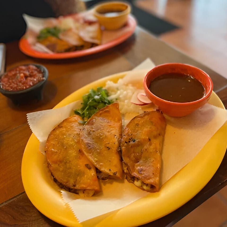 Three Krave Tacos birria tacos on a yellow plate with consommé, salsa, and cilantro.