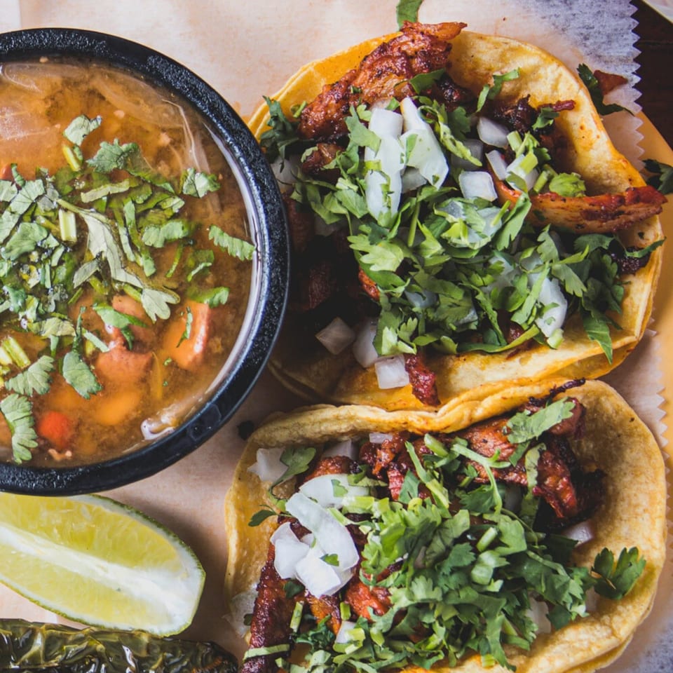 Two Krave Tacos with cilantro and onions, a bowl of birria, and a lime wedge.
