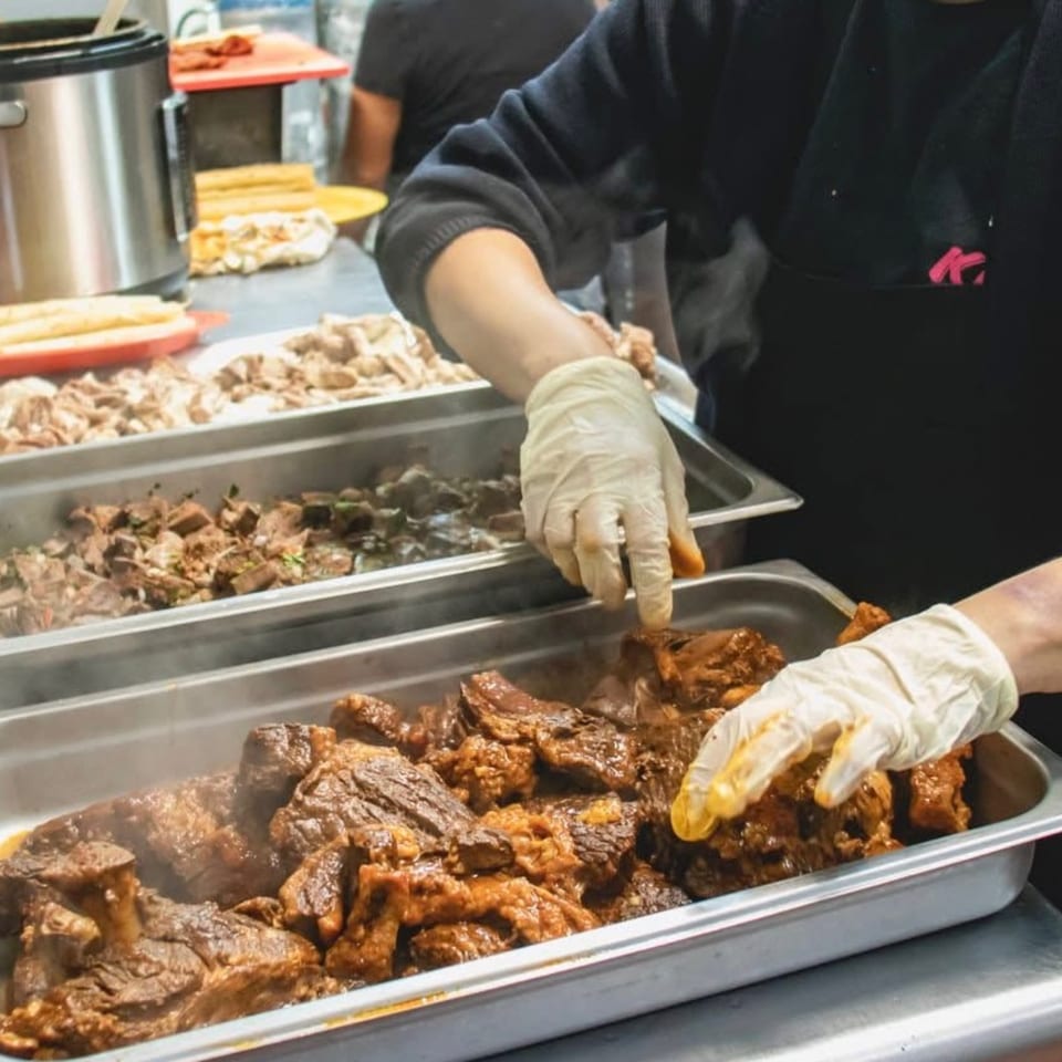 Gloved hands serving Krave Tacos' Mexican braised beef from a steaming buffet tray.