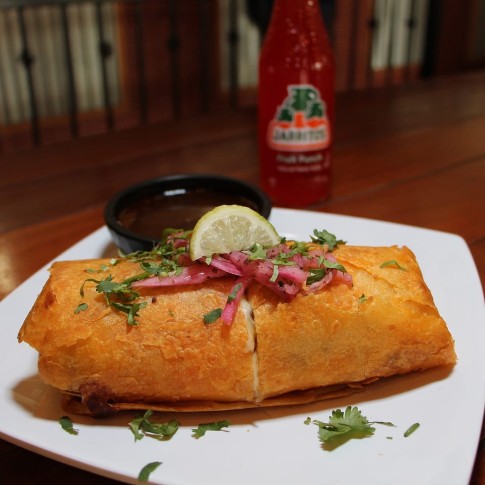 A golden-brown chimichanga topped with pickled red onions, cilantro, and a lime wedge, with a Jarritos soda in the background.