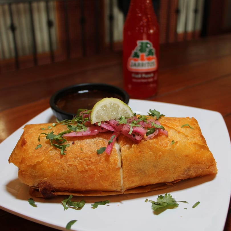 A golden-brown chimichanga topped with pickled red onions, cilantro, and a lime wedge, with a Jarritos soda in the background.