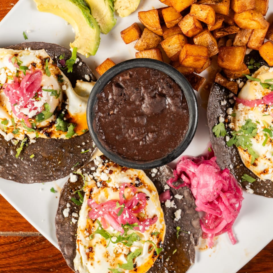 Three Krave Tacos with fried eggs, pickled onions, cilantro, and cheese, served with black beans, potatoes, and avocado.