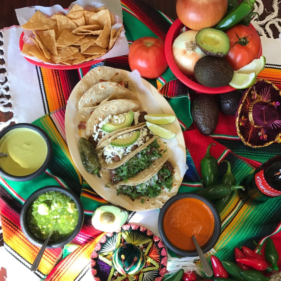 Overhead view of Krave Tacos' Mexican feast with tacos, chips, salsas, and fresh ingredients on a colorful serape.